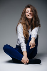 Portrait of young exited fashion girl sitting on floor and posing in casual clothes on grey