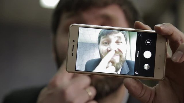 Busy office worker cranks and looking into phone camera. funy businessman picks finger in nose, strokes his beard