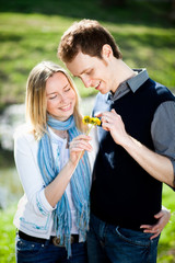 Tender couple in love with yellow dandelions flowers in the park in spring. Happy girl and guy smile and hug outdoor, they are wearing in jeans, scarf, vest.
