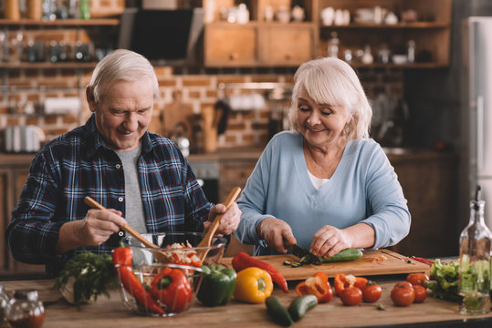 Portrait Of Senior Woman Cutting Cucumbers While Husband Mixing Vegetables For Salad