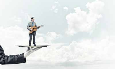 Businessman on metal tray playing acoustic guitar against blue sky background