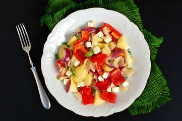 Salad with potatoes, red pepper, onion, basil and feta cheese on white plate on grey cloth on black...