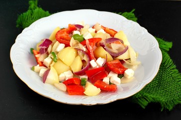 Salad with potatoes, red pepper, onion, basil and feta cheese on white plate on grey cloth on black background