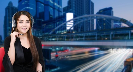 portrait of asian woman support phone operator or call center in headset sitting on red chair on blurred night city background, customer support and service concept.