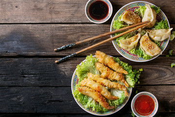 Fried tempura shrimps and gyozas potstickers on lettuce salad with sauces and rice. Served in traditional china plate with chopsticks over old wooden background. Top view. Asian dinner