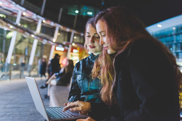 Two young beautiful caucasian women friends outdoor in the city night using computer, tapping keyboard - business, technology, social network concept