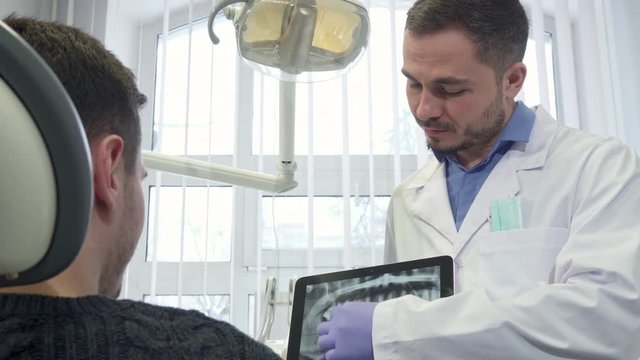 Middle aged male dentist showing brunette client the x-ray on his tablet. Bearded dental specialist zooming the image on his gadget. Close up of doctor in white coat in sterile gloves explaining