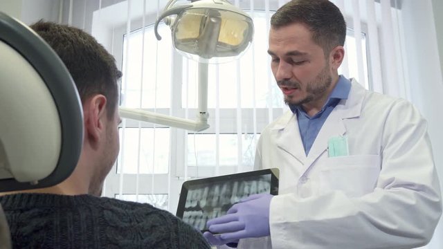 Caucasian dental specialist moving the image on the touchscreen. Brunette bearded doctor holding tablet in his hands. Close up of male patient sitting backwards on the chair