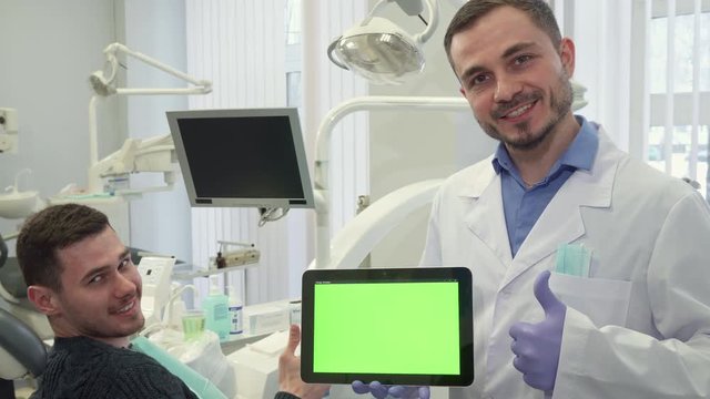 Middle aged dentist showing his thumb up at his office. Attractive bearded dental specialist holding digital tablet with green chroma key on it's screen. Caucasian male doctor in white coat standing