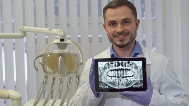 Middle aged bearded dentist demonstrating the x-ray of human teeth on his tablet. Brunette bearded dental specialist holding digital gadget in his hands at the office. Camera crabbing left to the male