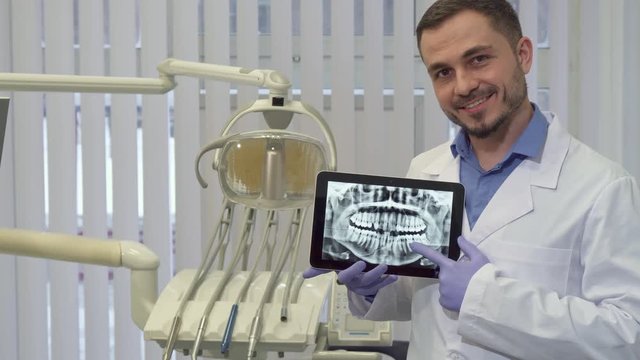 Handsome brunette dentist pointing his forefinger on the x-ray. Attractive caucasian doctor looking at the tablet screen. Middle aged dental specialist nodding at his office