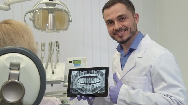 Attractive caucasian dentist approving tooth health on the x-ray. Middle aged brunette doctor gesturing ok to his female patient. Handsome bearded dental specialist looking into the camera with tablet