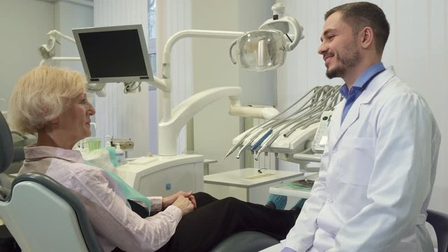 Middle aged handsome dentist greeting his female client at his office. Brunette bearded doctor shaking hands with senior blond woman. Attractive caucasian doctor in white coat and sterile gloves