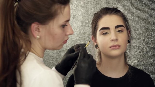 Makeup artist fixing the shape of painted eyebrows in beauty salon