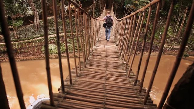 Tourist guy walks on a suspension bridge made of bamboo in the jungle.