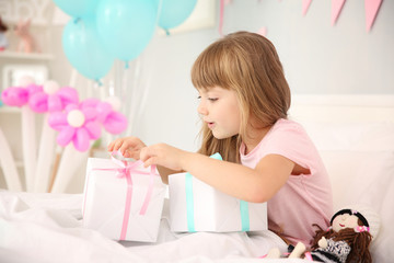 Cute birthday girl opening boxes with presents while sitting on bed at home