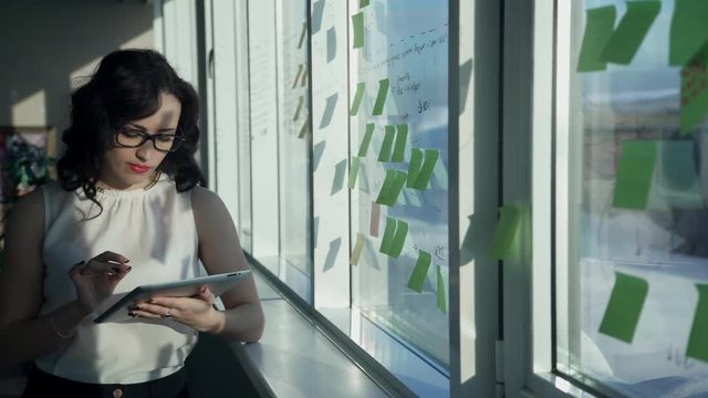 office worker woman looking to tablet, stickers pasted on window. Brunette in white shirt with long hair working with device, touches screen. Business female analyzing important topics, looks at notes