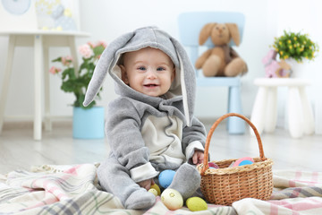 Naklejka premium Cute little baby in bunny costume playing with Easter eggs at home
