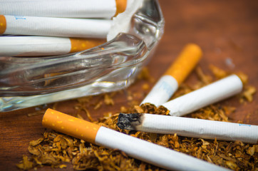 Closeup cigarettes and tobacco lying inside and around glass ash tray on wooden surface, anti...
