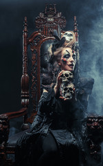 Fototapeta na wymiar Young beautiful witch sits on a chair. Bright make up, skull, smoke- halloween theme.