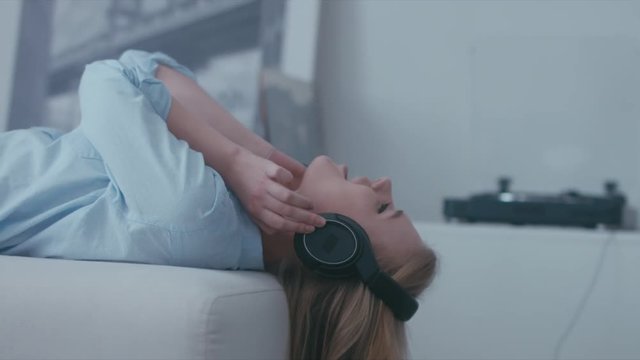 Young Caucasian teen girl lying on a sofa at home with headphones on and singing while listening to music from a vinyl turntable. 4K UHD RAW edited footage