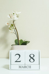 Closeup white wooden calendar with black 28 march word with white orchid flower on white wood desk and cream color wallpaper in room textured background , selective focus at the calendar