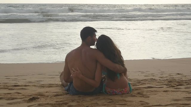 Young romantic couple is enjoying beautiful view sitting on the beach and hugging at evening. A woman and a man sits together in the sand on the sea shore, admiring the ocean and landscapes.Slowmotion