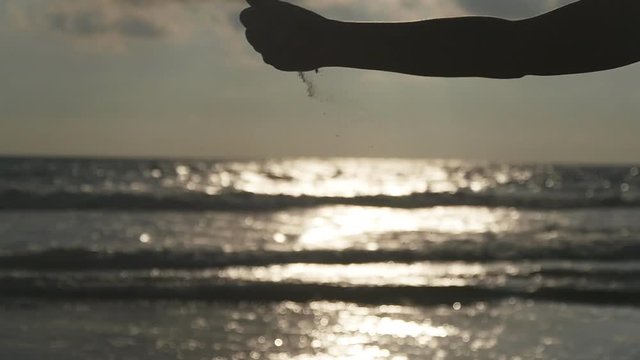 Female hand pouring sea sand through her fingers at sunset against an ocean background. Arm of young woman with sand strewed or falling from it. Grit drizzling from fist of girl. Slow motion Close up