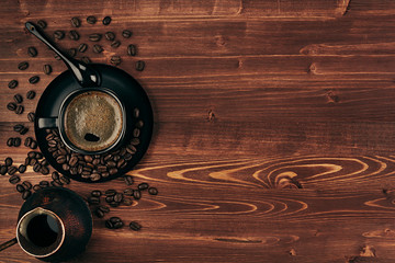 Hot coffee in black cup with beans, spoon and turkish pot cezve with copy space on brown old wooden board background, top view.  Rustic style.