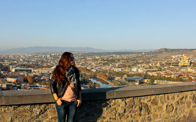 Fototapeta premium Girl in the sunglass looks. Panoramic view of old Tbilisi, Georgia at the background. Traveler woman walking around the city by looking at the view from above. The view from the castle Narikala.