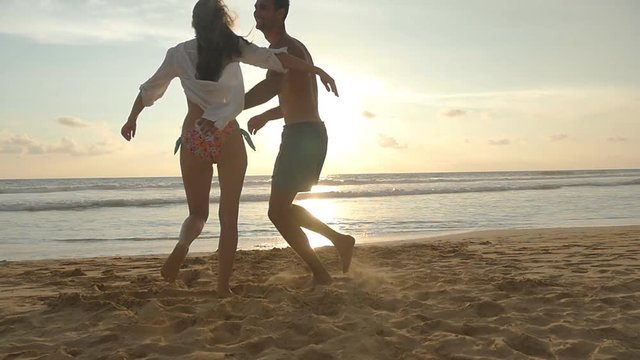 Young couple is running on the beach, man hug and spin around his woman on sunset. Girl jumps into her boyfriend arms, he whirling her at beautiful seaside. Having fun together at vacation. Slowmotion
