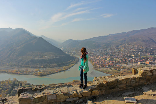 Traveler positive girl standing on the stone wall. Territory Jvari Monastery. City Mtskheta, rivers Aragvi and Kura, and stunning mountains at the background. Freedom concept.
