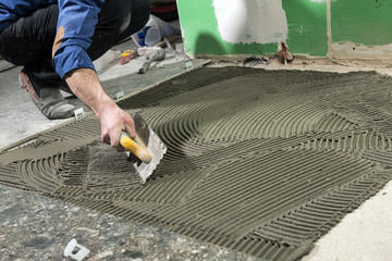 Real construction worker applying cement adhesive for ceramic tile on floor.