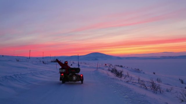Two motorcycles with sidecars ride along snow-covered road far beyond Polar Circle against backdrop of scarlet sunset