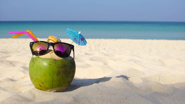 Fresh coconut with drinking straw and sunglasses on sandy white beach. Dolly shot UHD
