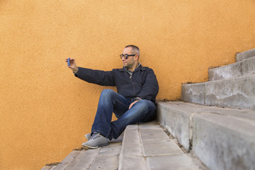 happy handsome man taking a picture with his smart phone over orange background. He is wearing modern sunglasses. Copy space. Lifestyle