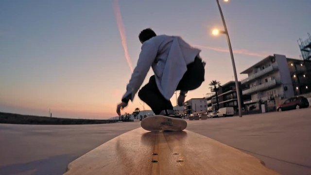 Dynamic action shot of cool unrecognizable guy jumping on long longboard from bamboo wood and surf it through beach promenade aggressively at beautiful sunset time