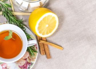 Cup of black or red tea with lemon, rosemary, mint, cinnamon sticks, sugar on grey tablecloth, top view