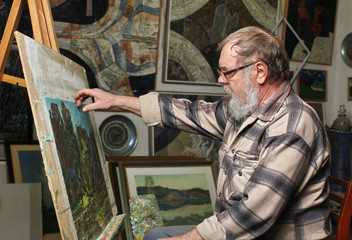Fototapeta premium Elderly painter with beard and glasses draws a flowers picture by oil paint in art workshop