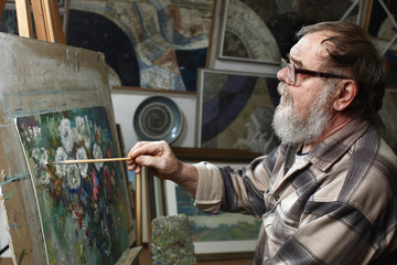 Elderly painter with beard and glasses draws a flowers picture by oil paint in art workshop