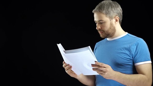 Handsome man in blue tshirt looking through papers. Cotract, bills, letter concepts. Black background. 4K video