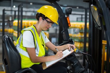 Female factory worker sitting on forklift and writing in clipboard at factory