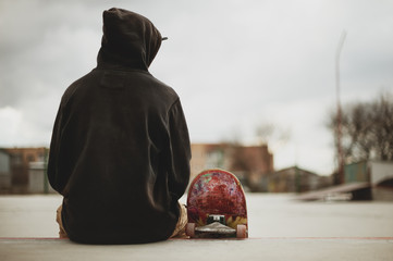 Teenager sitting in a black sweatshirt holding a skateboard on a slum background urban © yanik88