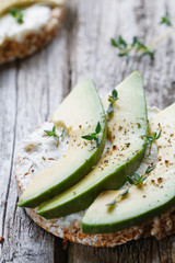 Close up of a toast of grain bread with soft white cheese and chopped fresh avocado on old wooden gray boards .