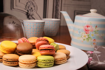 Teapots and cups on a wooden table. White plate and macaroon with white glass case. Homemade pastries. Cozy atmosphere.
