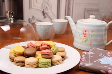 Teapots and cups on a wooden table. White plate and macaroon with white glass case. Homemade pastries. Cozy atmosphere.