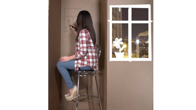 Tiny apartment in a cardboard box woman drawing flower on a wall. Painted window with a view of the night city and high-rise apartment building