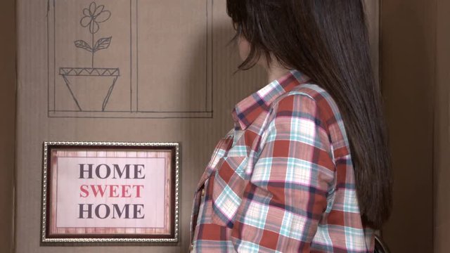 Woman living in a cardboard box with HOME SWEET HOME picture frame on the wall of her tiny apartment