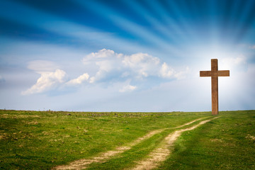 Jesus Christ cross, wooden crucifix on a scene with blue spring sky, green meadows, bright light, rays, clouds. Christian wooden cross on field with green grass and path leading to the cross