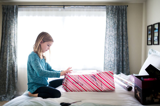Side view of girl wrapping gift box while kneeling on bed